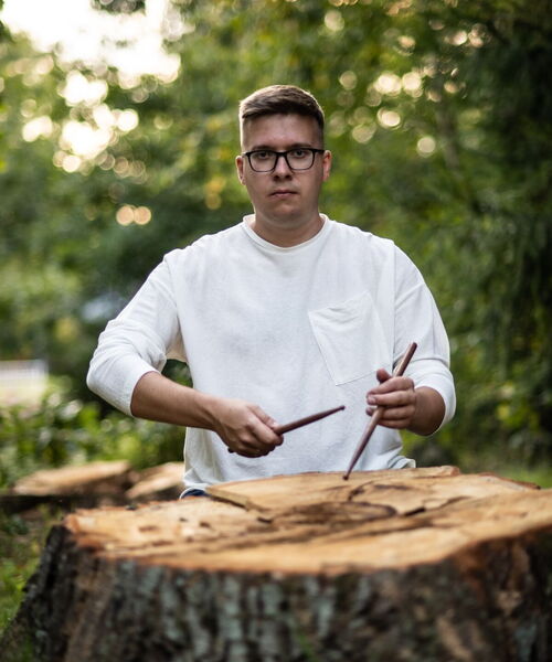 Zu sehen ist Rostislav Balciunas vor einem abgesägten Baum mit Drumsticks in der Hand.